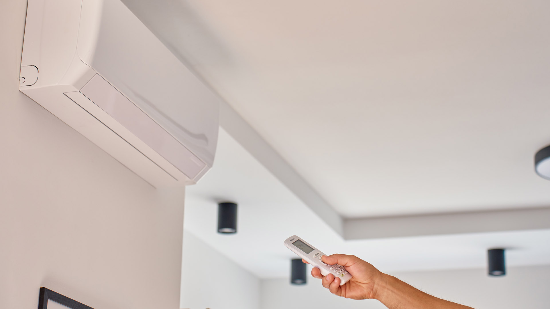 Klimatyzator ścienny zamontowany na białej ścianie, a w dłoni widoczny pilot sterujący urządzeniem.