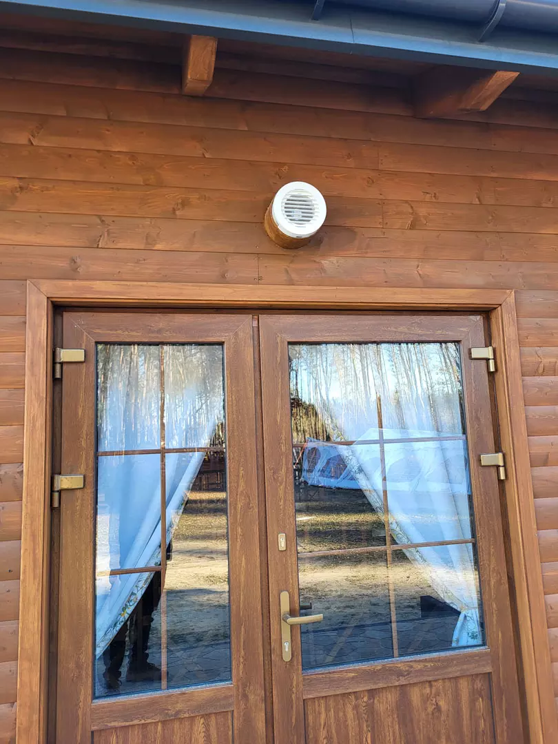 Dwuskrzydłowe drewniane drzwi z szybami i zasłonkami oraz biała kratka wentylacyjna zamontowana na drewnianej elewacji nad drzwiami.