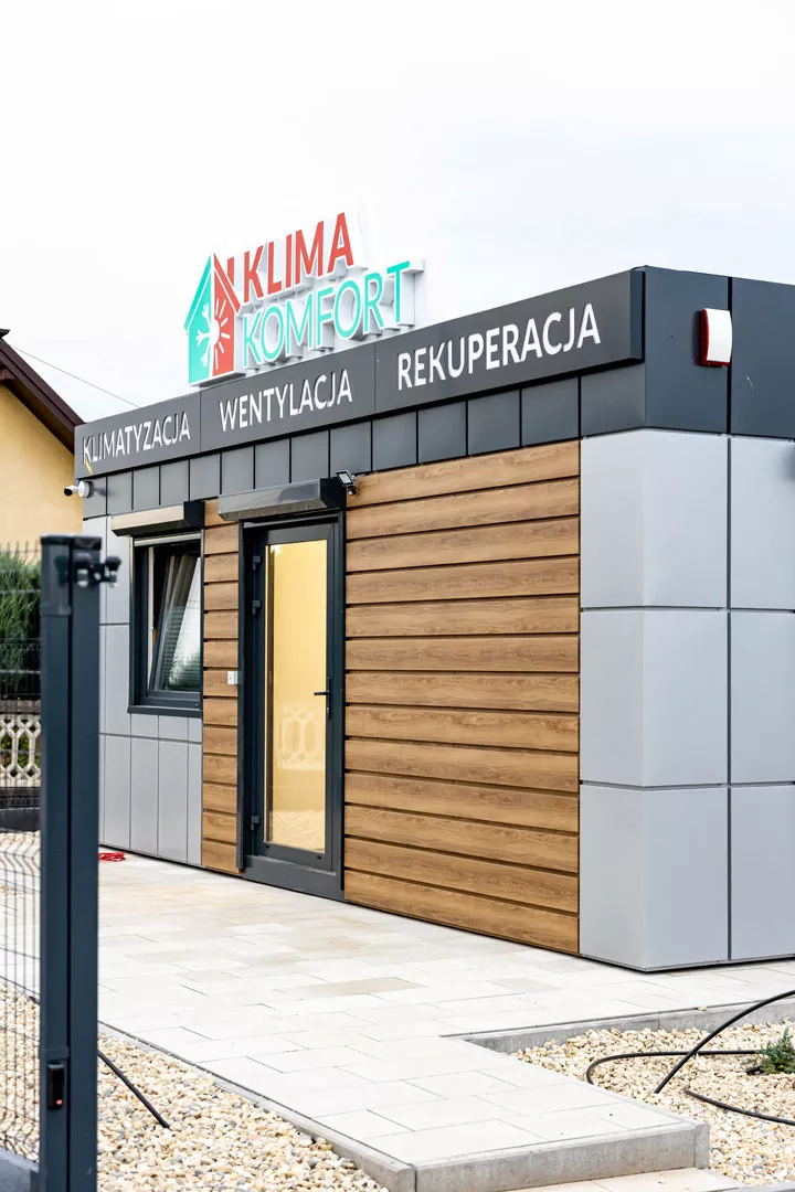 Nowoczesna siedziba firmy Klima Komfort z podświetlonym logo i napisami „Klimatyzacja, Wentylacja, Rekuperacja” na elewacji, drewnianymi panelami i szklanymi drzwiami.