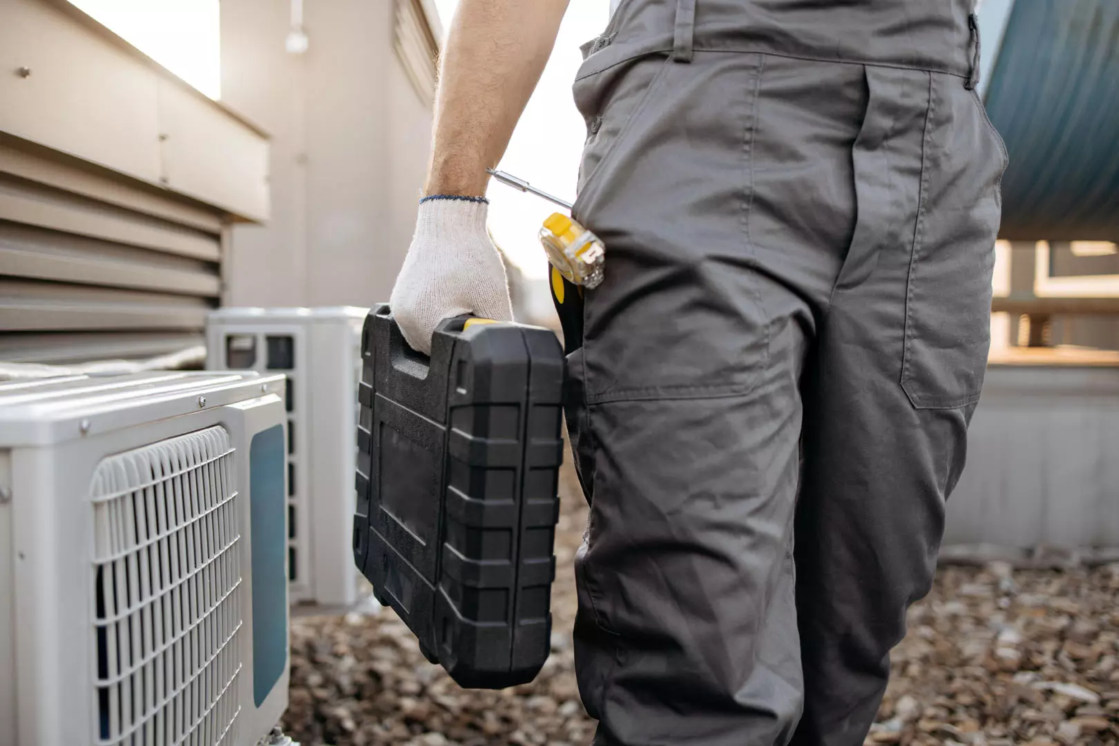 Technik serwisujący klimatyzację w roboczym ubraniu trzyma skrzynkę narzędziową obok zewnętrznych jednostek klimatyzacyjnych.